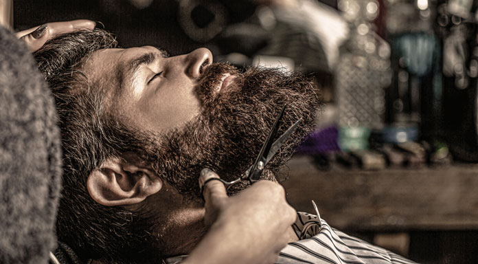 Körperpflege - Mann sitzt beim Babier auf dem Frisierstuhl und lässt sich den Bart trimmen.