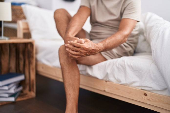 Körperpflege - Mann sitzt auf der Bettkante und hält sich den linken Fuß fest.