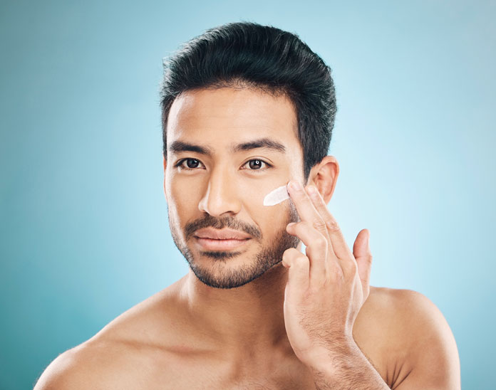 Körperpflege - Gutaussehender Mann steht vor dem Spiegel und trägt sich eine Hautcreme auf sein Gesicht auf.
